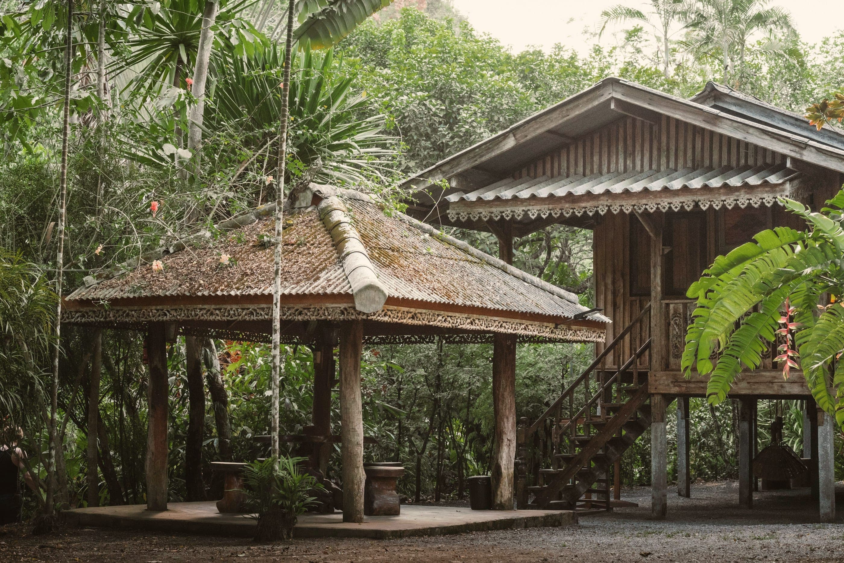 A wooden house on stilts beside an open carved pavilion, tall trees behind.
