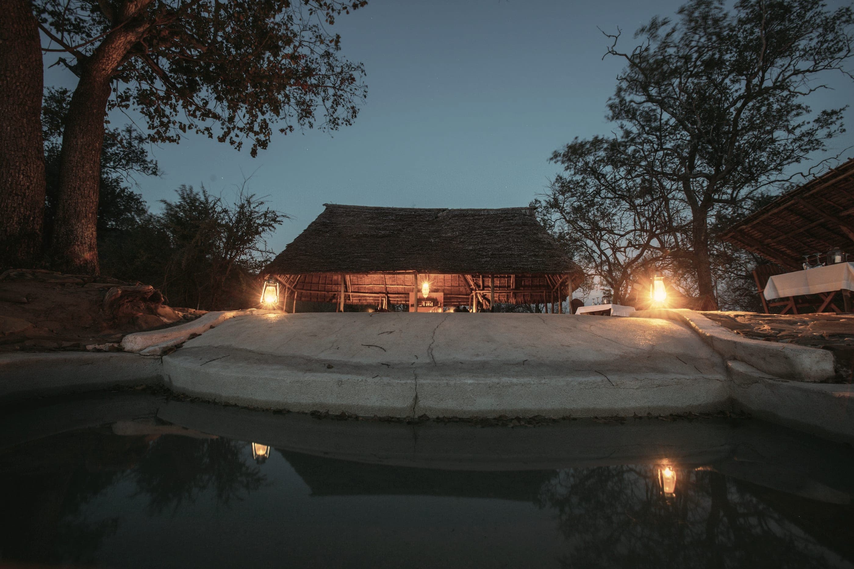 A thatched lodge at dusk, two lamps lit, the pool holding their light.