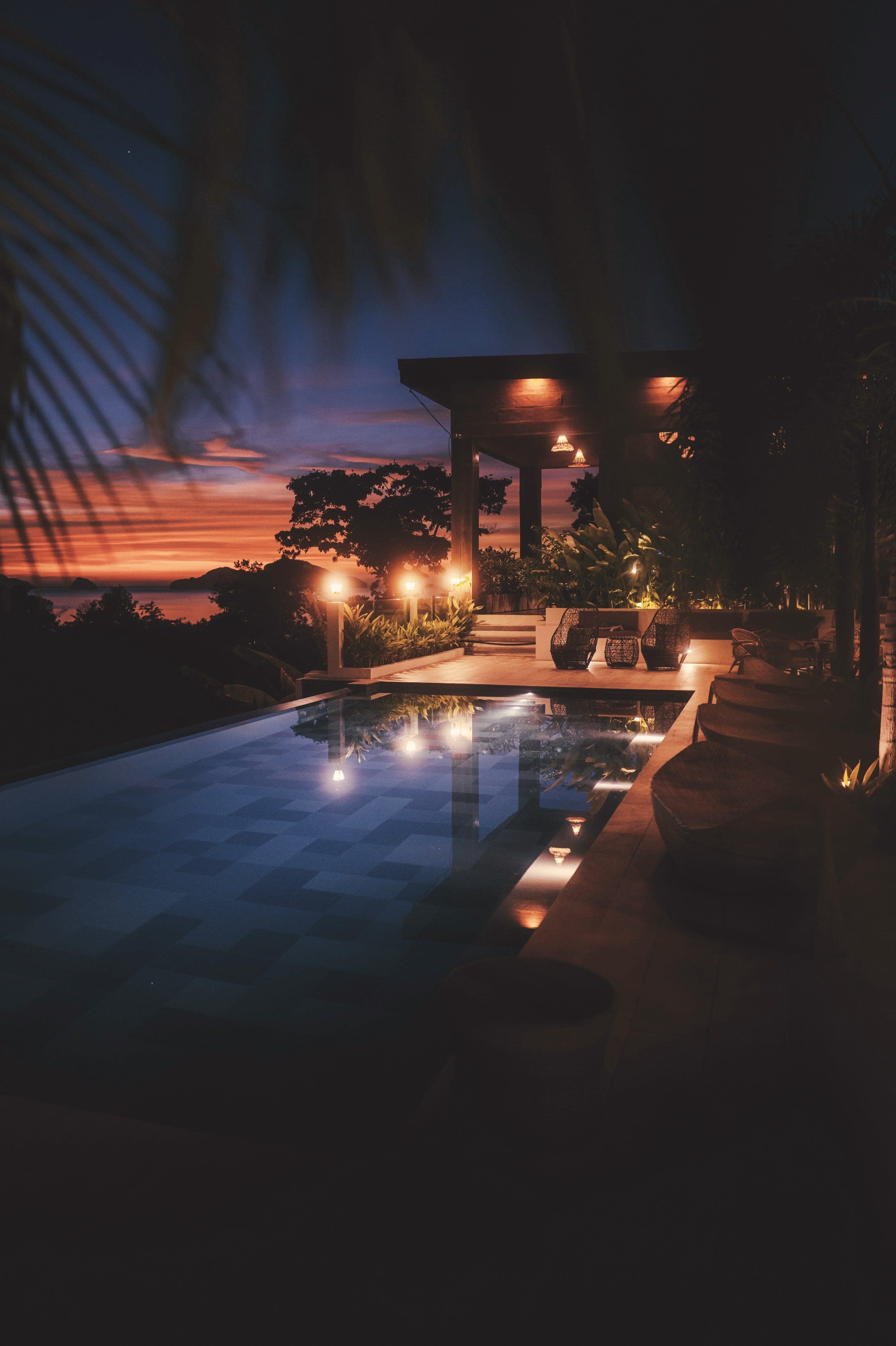 A pool lit from under the water at night, lamps warm along the deck, the last of the sunset behind.